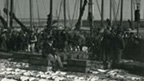Lines of large fish are displayed for sale on the ground. Various people stand around, with a line of fishing boats and the harbour lighthouse behind.