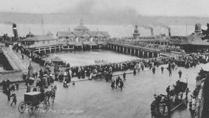 Black and white view of Dunoon Pier lined with crowds of people. Horses and carriages are on the street in the foreground. Two steamers with smoking chimneys are moored in the background.
