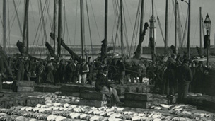 Lines of large fish are displayed for sale on the ground. Various people stand around, with a line of fishing boats and the harbour lighthouse behind.