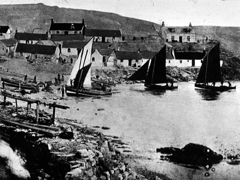 Black and white view of small sailing boats landing on a beach. A cluster of cottages rises up the hill behind.