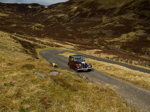 Colour view of a vintage Daimler motorcar climbing a straight section of road with a hairpin bend behind. The surrounding mountainous area is covered in grass and heather.