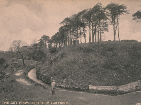 A small figure stands on a footpath by a narrow man-made water course at the foot of a hill with trees around.