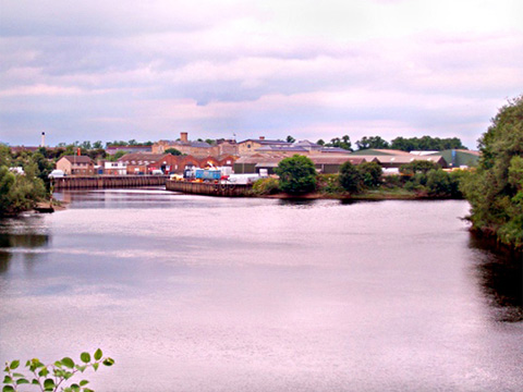Colour view of Perth Harbour from the south. The harbour side features some low warehouses and metal containers. In the background are some large, possibly industrial buildings.