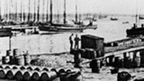 Black and white view of Baltasound Harbour. The quayside is covered with many rows of barrels. A number of single and two-masted boats are moored in the background.