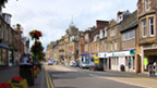 Colour view of Crieff High Street. The street is largely empty, save for a few parked cars and a van driving off into the distance. To the left of frame, the street is lined with ground level and raised planters containing red flowers and foliage 1