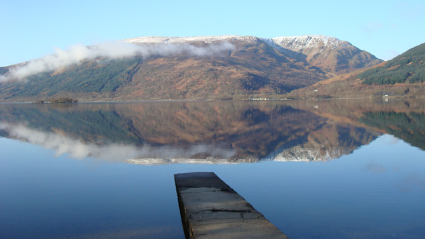 A view across Loch Lomond, with snow dusted hills and light cloud in the distance.