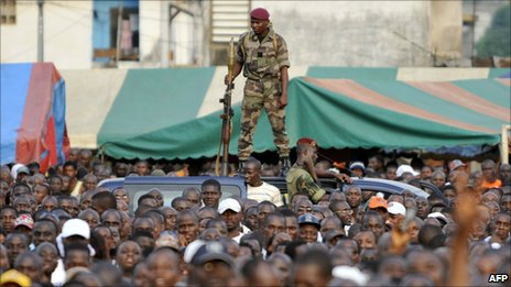 Photo showing a crowded street with a soldier holding a gun standing on top of a van