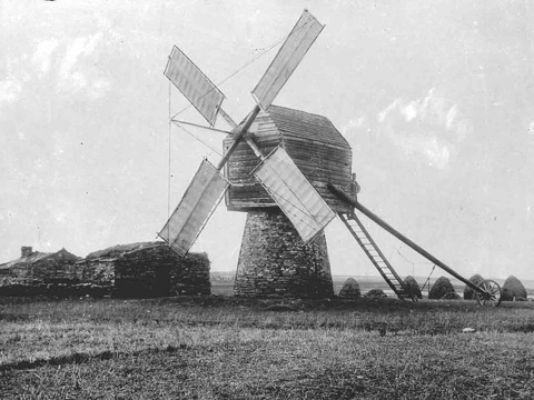 Four sailed timber and stone construction windmill with low stone buildings behind.