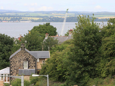 Colour view over rooftops, established trees and the top of a crane to wide river beyond.