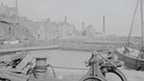 Black and white view of a harbour in which three tall-masted sailing ships are docked. In the foreground are a winch and scattered pieces of dockyard equipment. A village featuring traditional stone buildings, a tollbooth and several tall, industrial chimney stacks, rises up a hill behind the harbour.