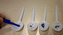 A pen-lid is used to drop some water into a spoons. 