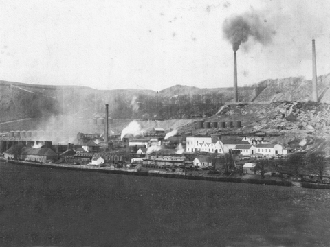 Black and white view of extensive industrial buildings with tall smoking chimney stacks with tall, flat topped hills behind.