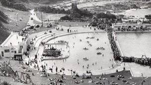 Black and white view, looking down from coastal cliffs to twin open air pools, with an Art Deco pavilion to the left of frame. One pool contains a mix of people paddling and rowing boats. The pool to the right is surrounded by a crowd of people, possibly watching a swimming competition. A large number of people are sunbathing on the surrounding grassy area.