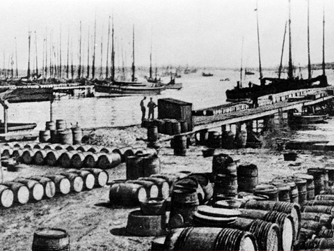 Black and white view of Baltasound Harbour. The quayside is covered with many rows of barrels. A number of single and two-masted boats are moored in the background.