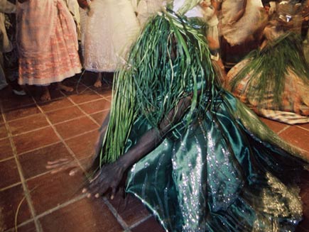 A man crouched on the floor slowly rises. He is wearing dark green palm fronds on his head, which completely cover his face. 
